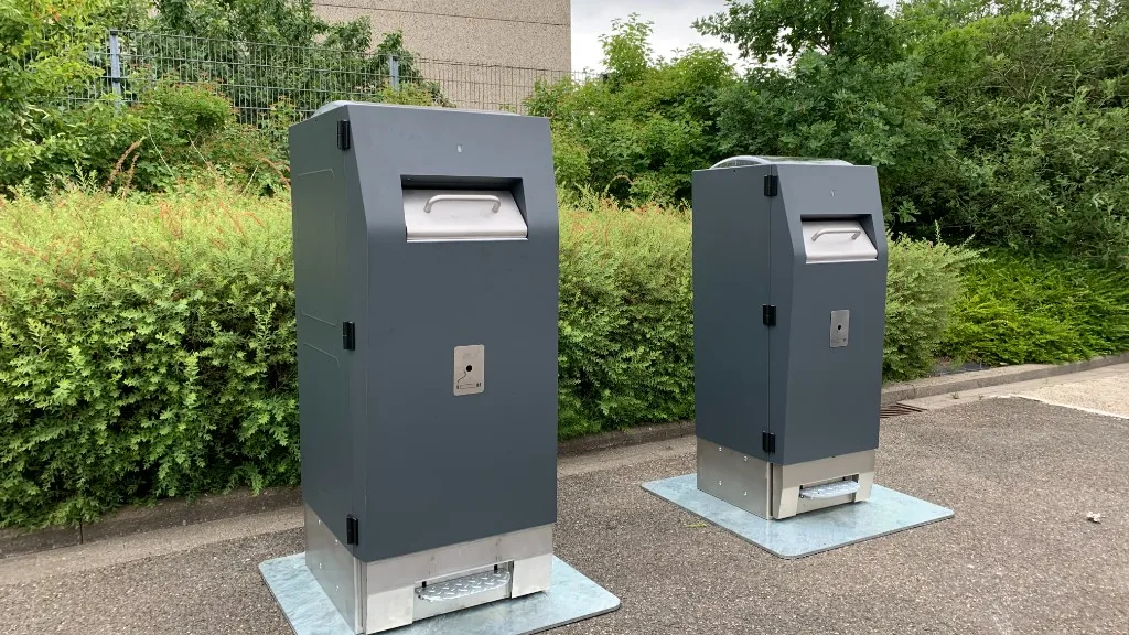 Due cestini WasteMate su area pedonale con siepe e alberi sullo sfondo